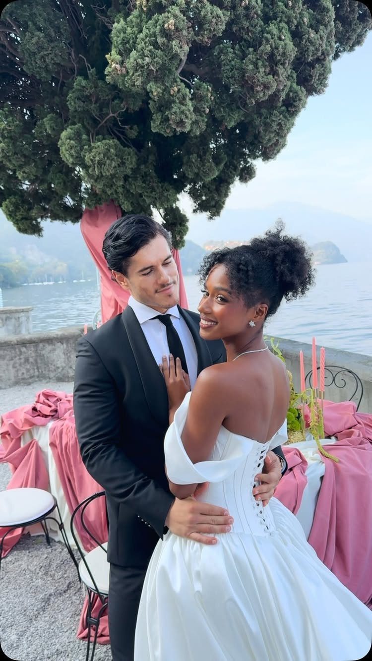 Couple in wedding attire posing by a lake with pink decorative fabric accents.