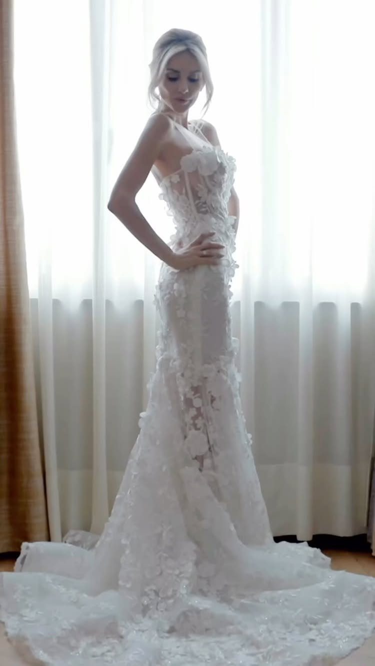 Woman in a white floral lace wedding dress posing against white curtains.