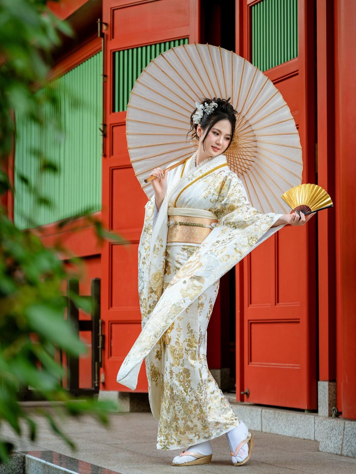 Woman in white and gold wedding kimono holding a parasol by red building