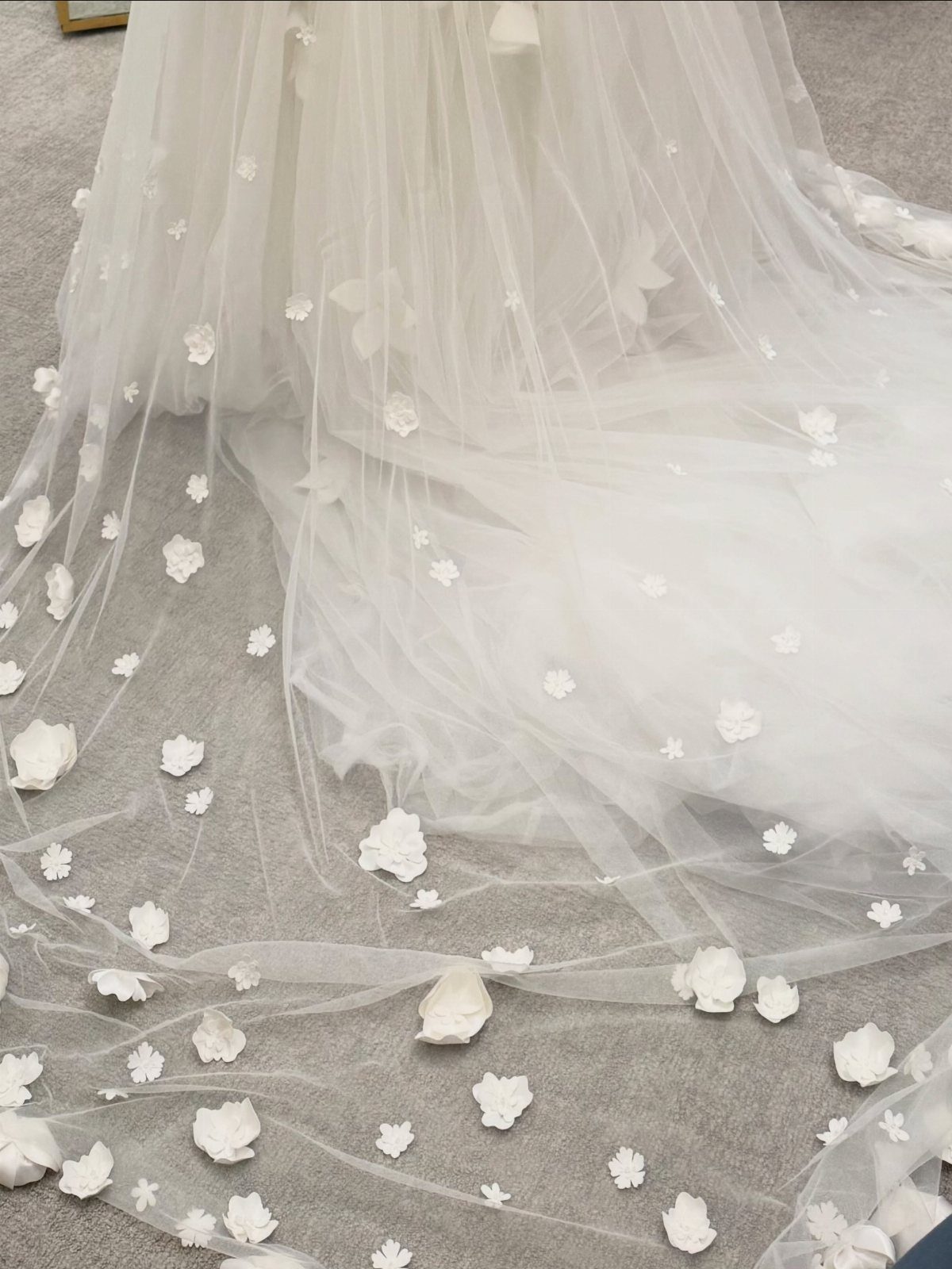 White wedding dress train with floral appliqués resting on a grey carpeted floor.