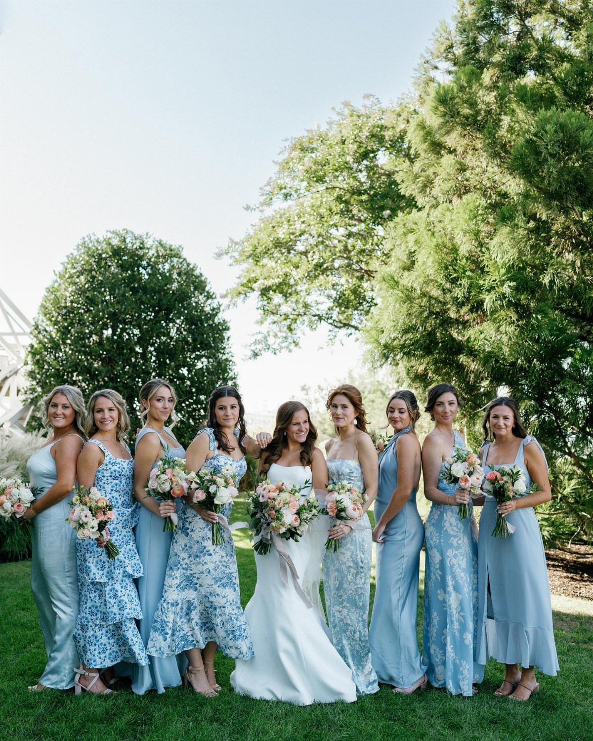 Bridesmaids in various blue dresses of different lengths standing with a bride outdoors.