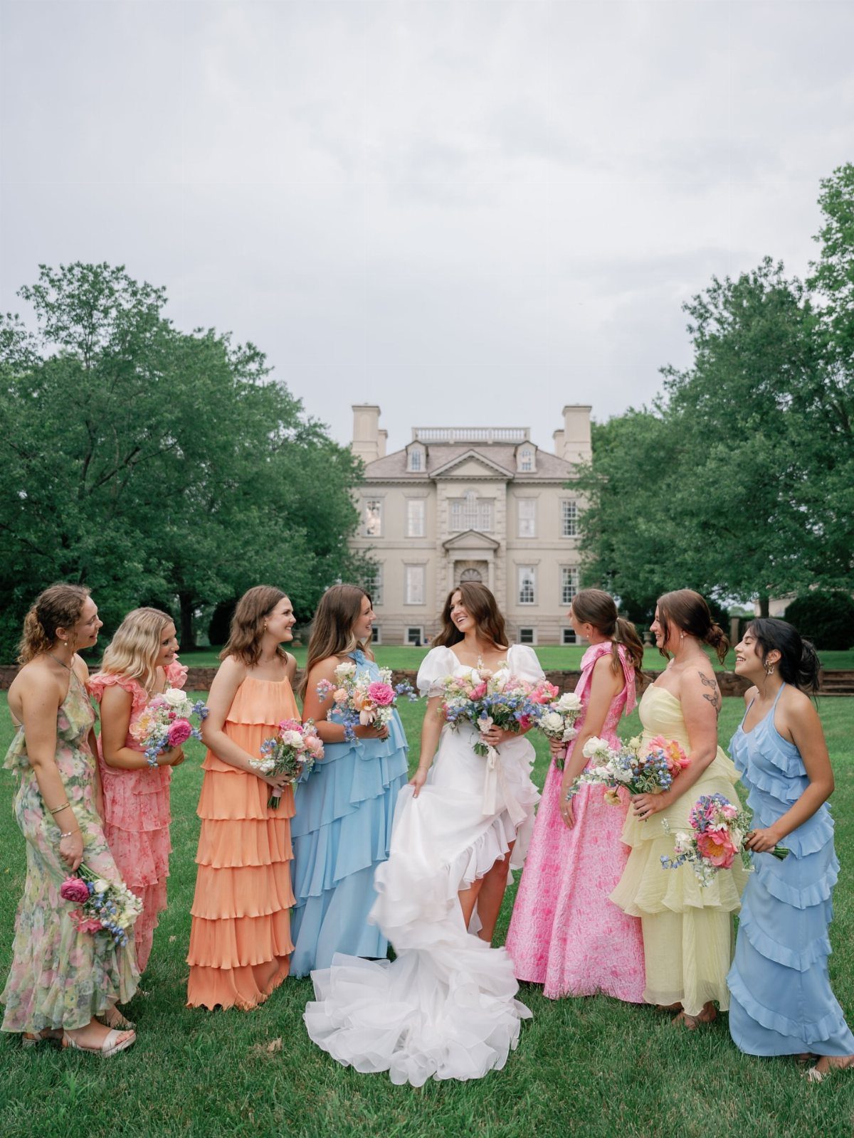 Bride and bridesmaids wearing rainbow colored mismatched dresses standing before a white manor.
