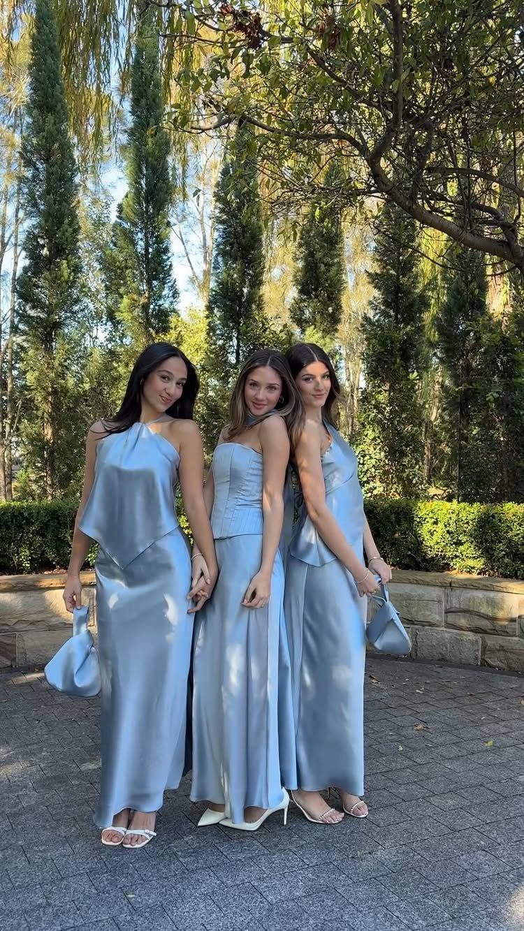 Three women wearing different styles of light blue satin bridesmaid dresses in a garden.