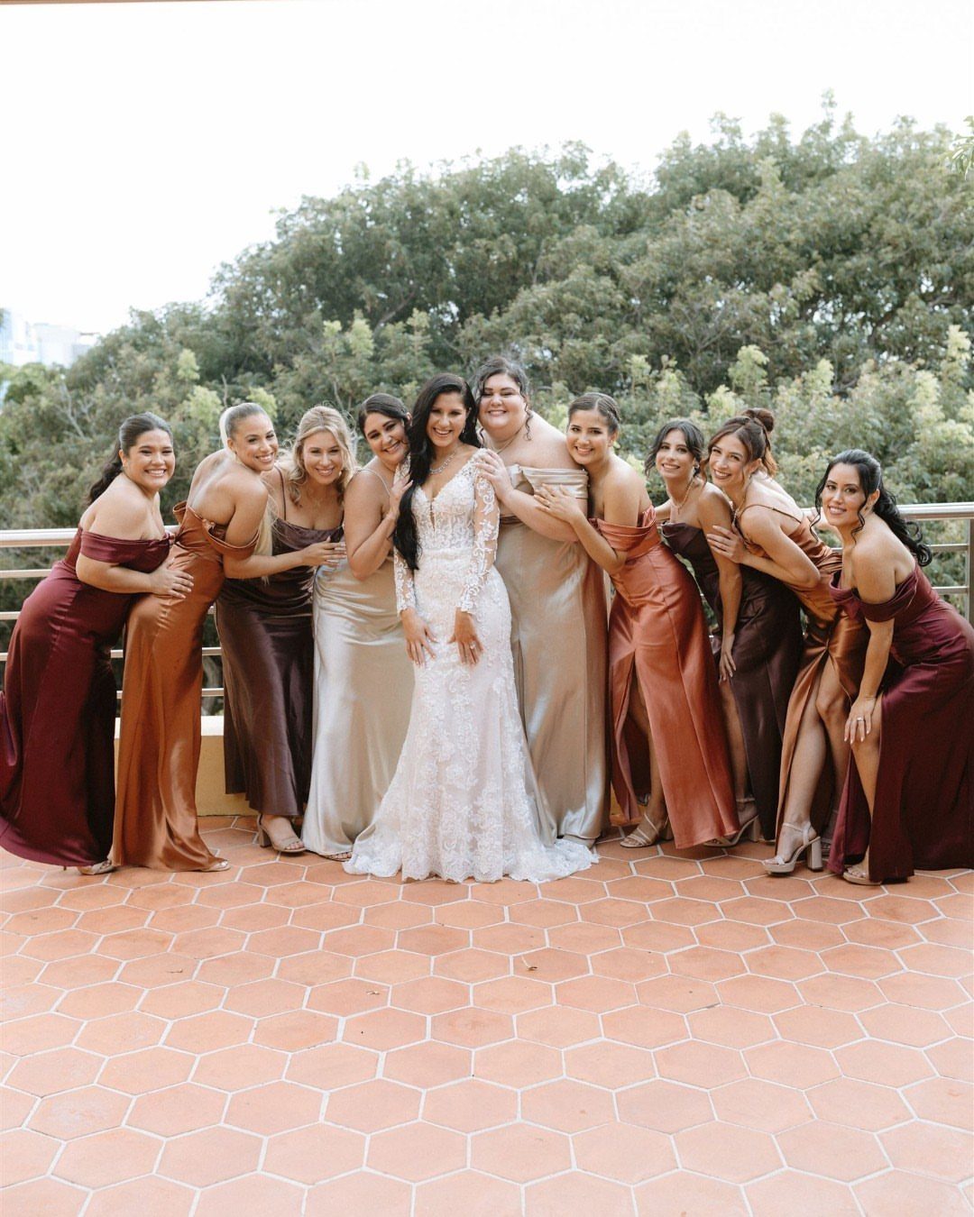 Bride in white lace gown with bridesmaids wearing mismatched satin dresses in earthy tones.