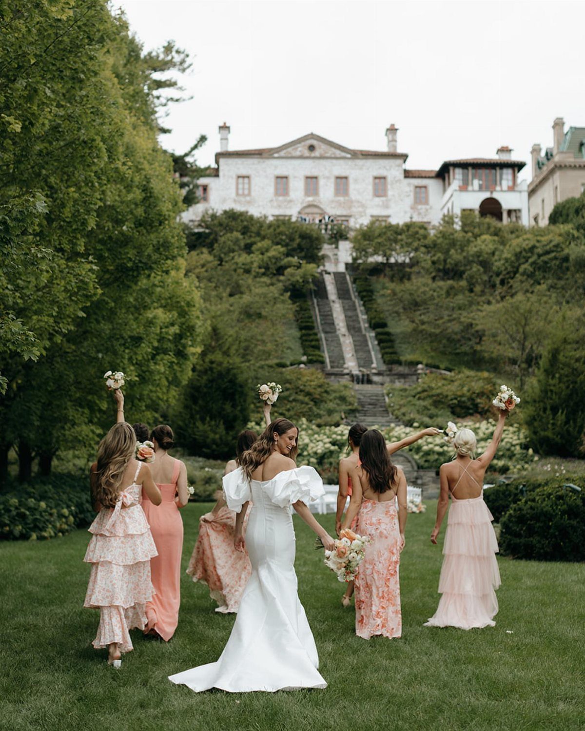 Bride and bridesmaids in mismatched pink and peach dresses on a lush green lawn.