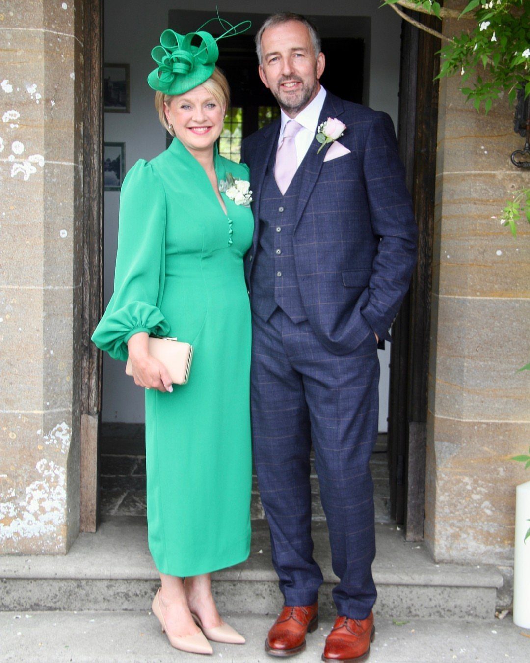 Woman in green midi dress and fascinator standing with man in navy suit.