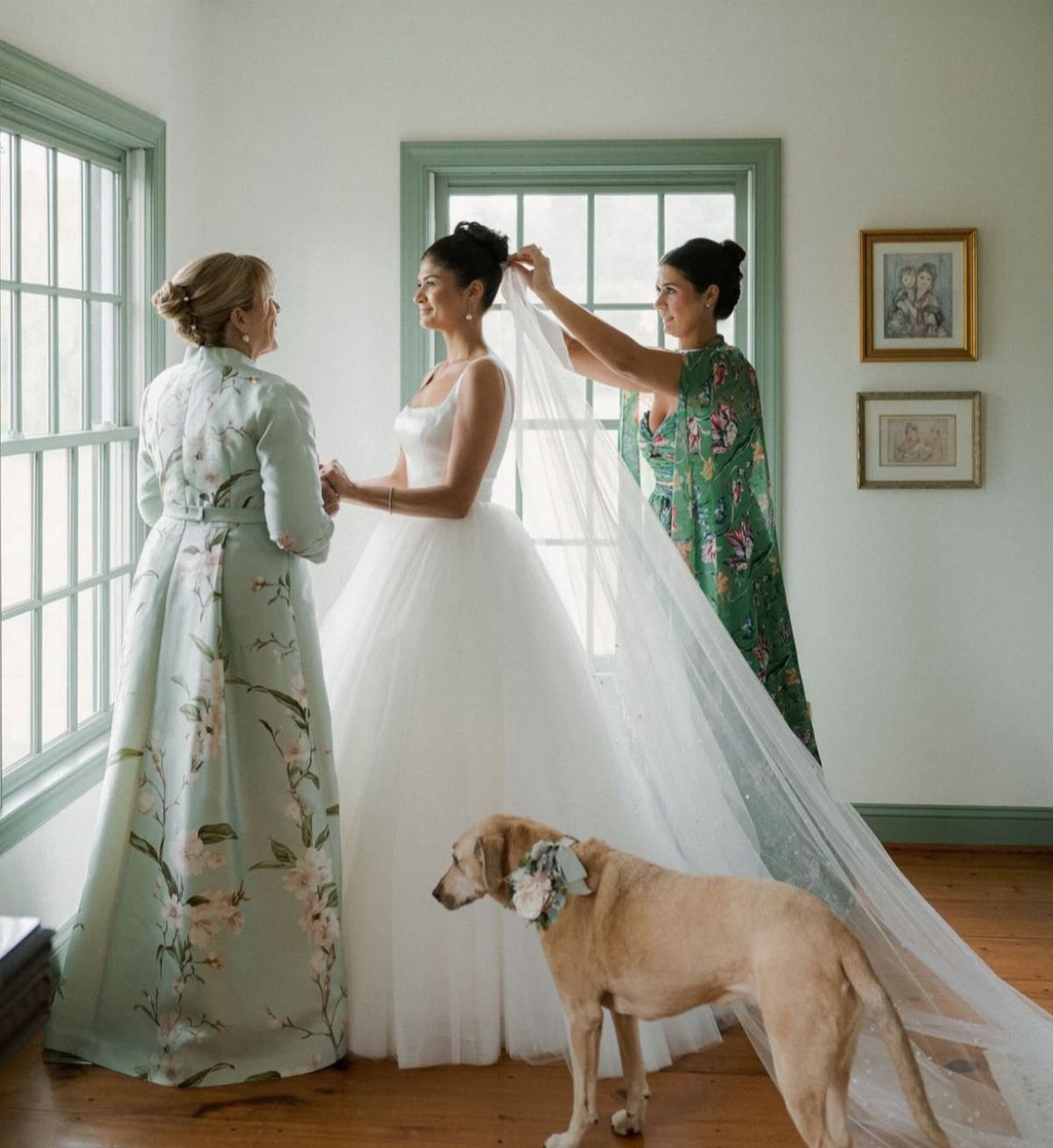 Two women in floral printed dresses assisting a bride in a white gown.