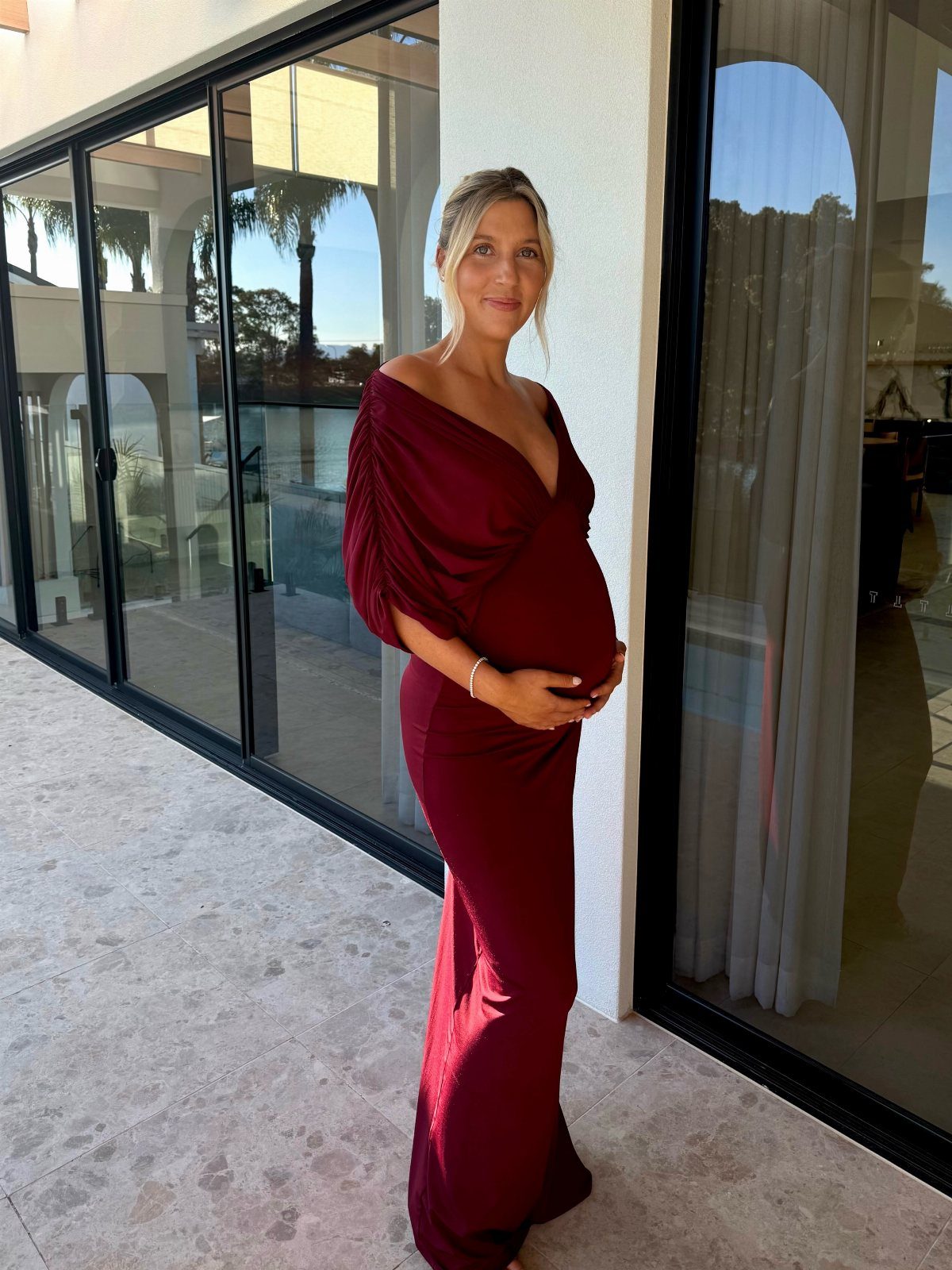 Pregnant woman in a long burgundy off-the-shoulder maternity dress standing on a patio.