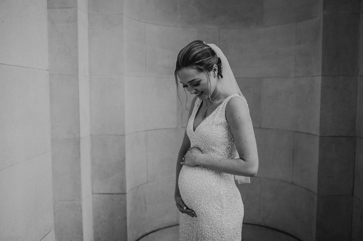 Pregnant woman in a textured white wedding dress and veil posing by stone wall