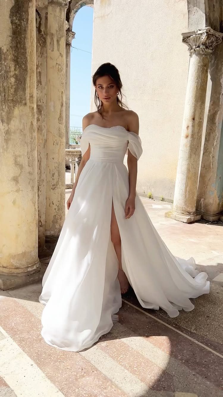 Woman in a white off-the-shoulder wedding dress standing between ancient stone columns.
