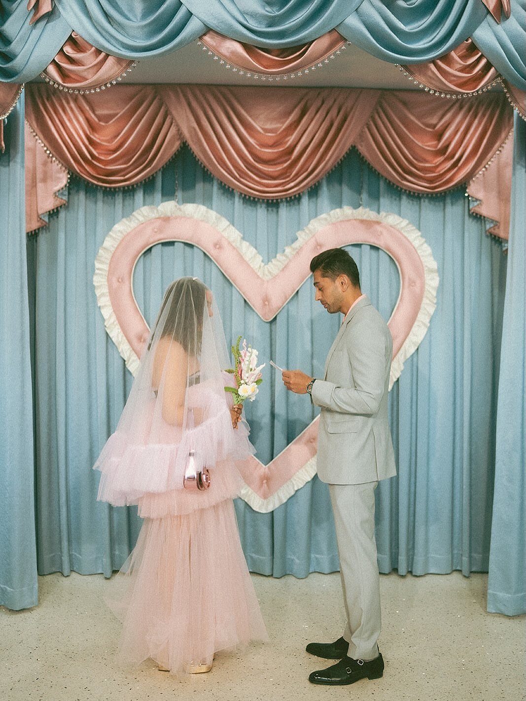 Bride in a pink tulle wedding dress and groom in grey suit facing each other.