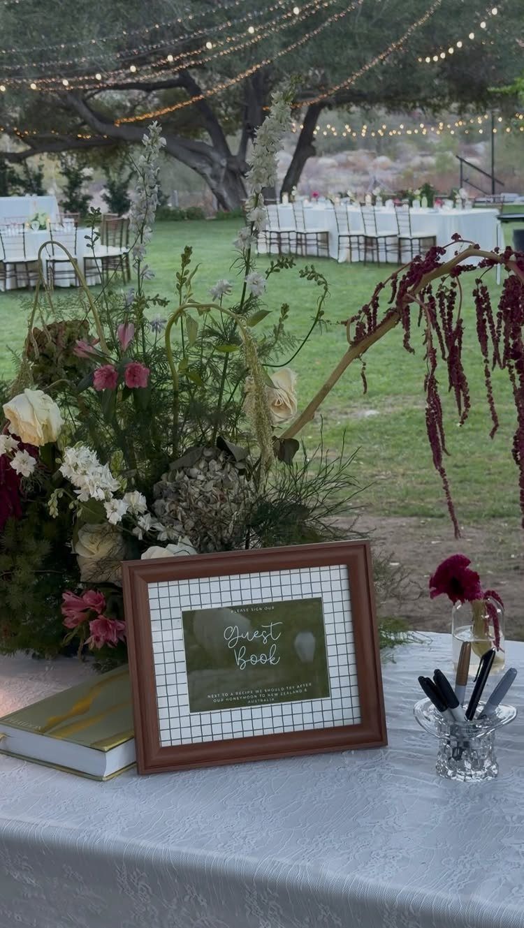 Guest book station with a framed sign, gold book, and floral arrangement outdoors.