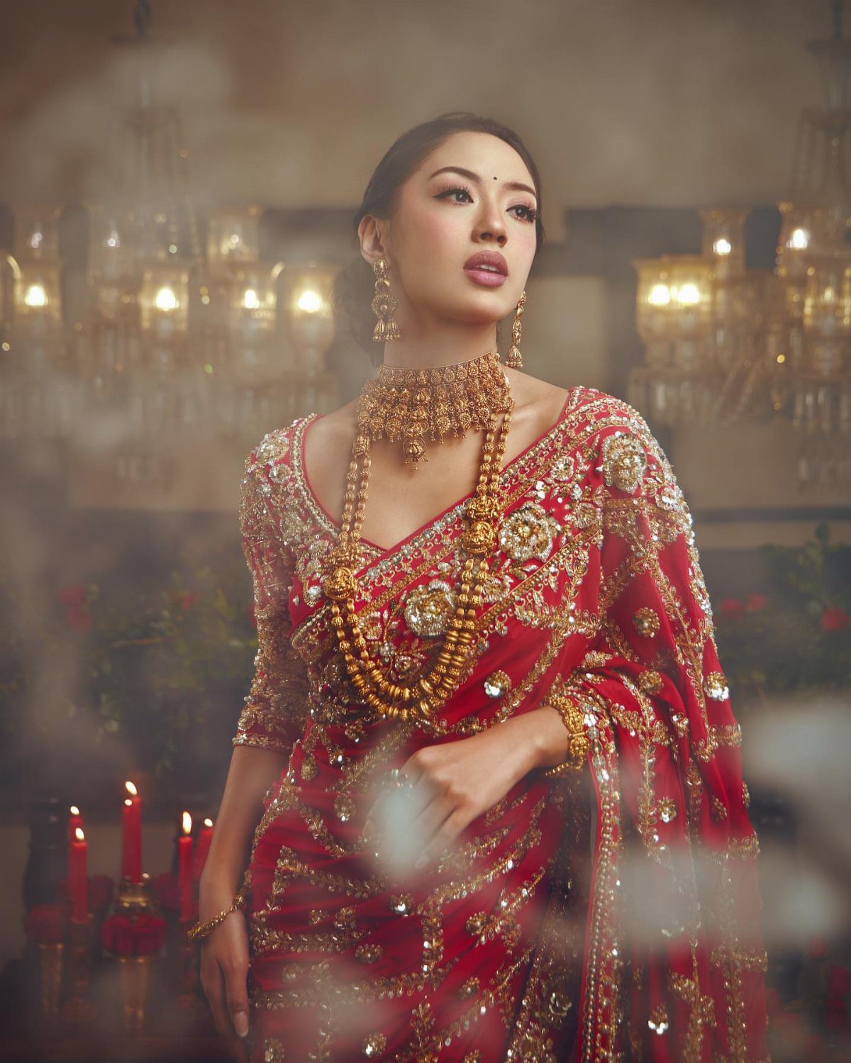 Woman in red embroidered Indian bridal lehenga with heavy gold jewelry and candles.
