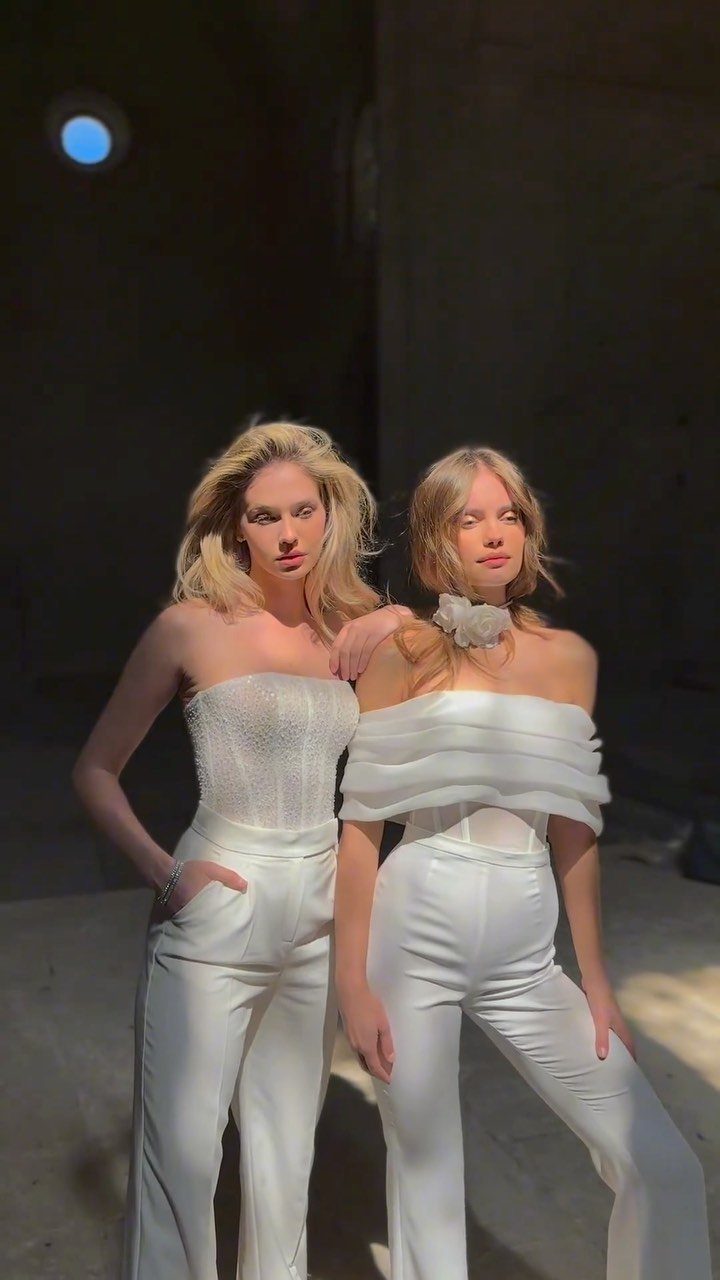 Two women wearing elegant white wide-leg jumpsuits posing in a concrete industrial space.