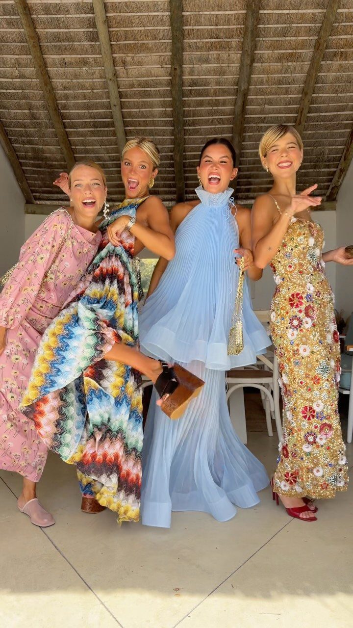 Four women wearing colorful floral and pleated dresses posing under a thatched roof.