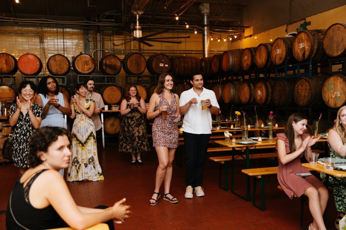 People in casual summer dresses and shirts gathering in a rustic winery setting.