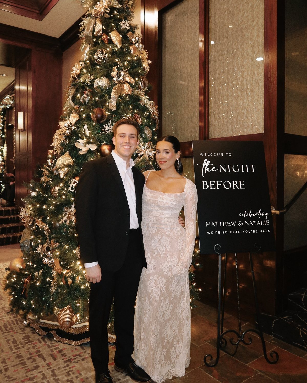 Couple in formal attire posing by a Christmas tree and welcome sign.