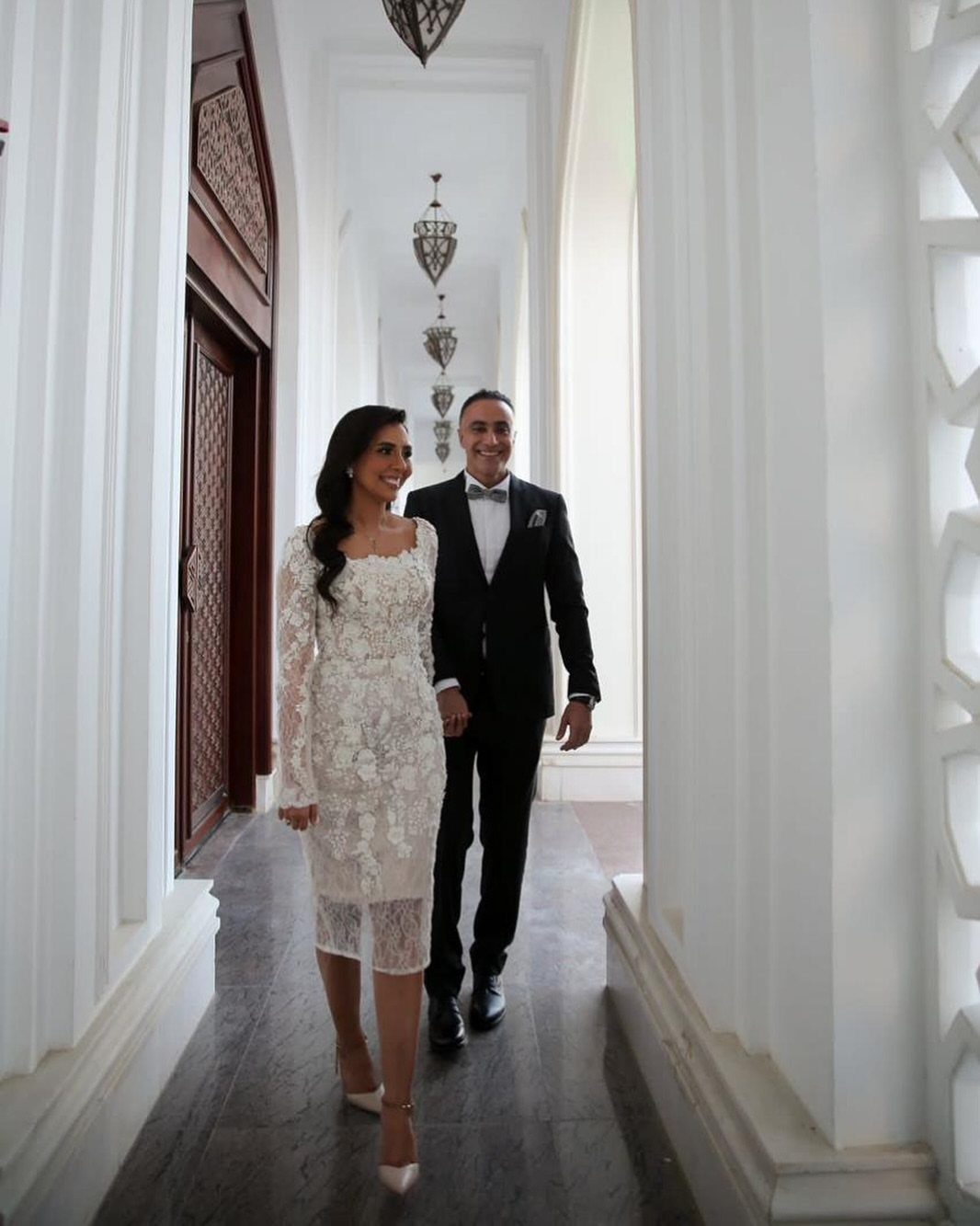 Woman in white lace midi dress and man in tuxedo walking in hallway