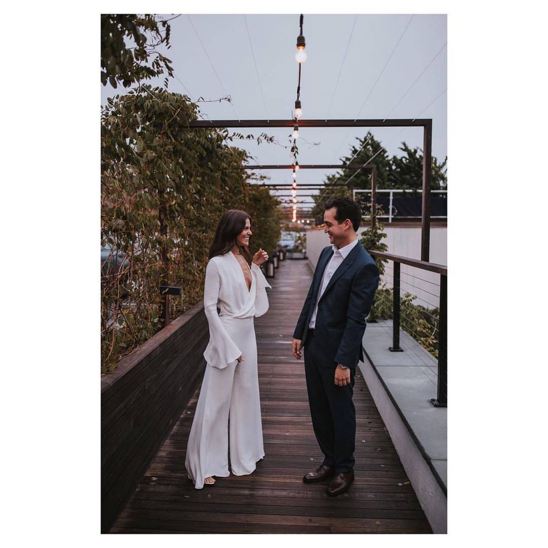 Woman in white wide-leg jumpsuit and man in blue suit on wooden deck