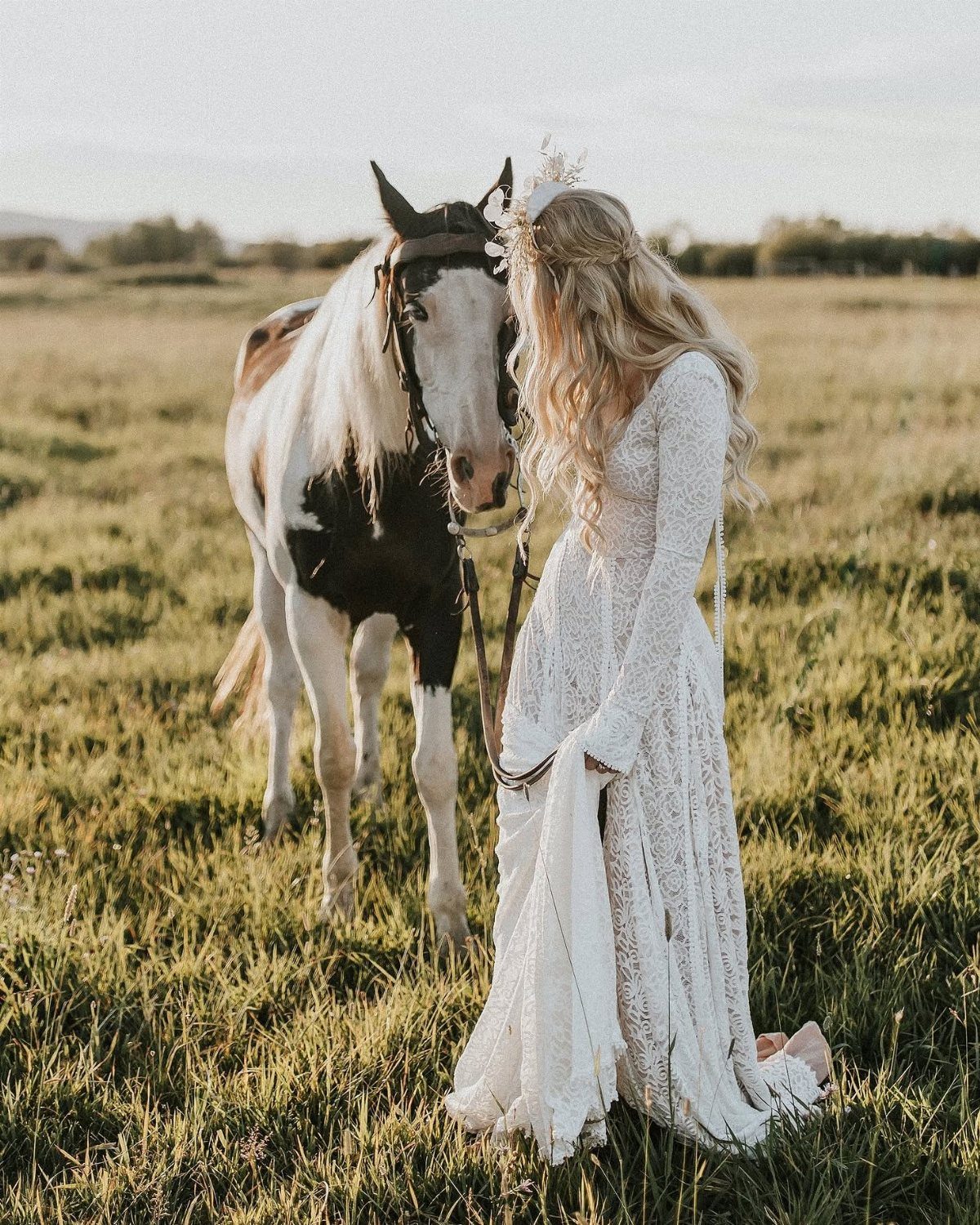 Woman in white lace wedding dress standing beside a horse in a field