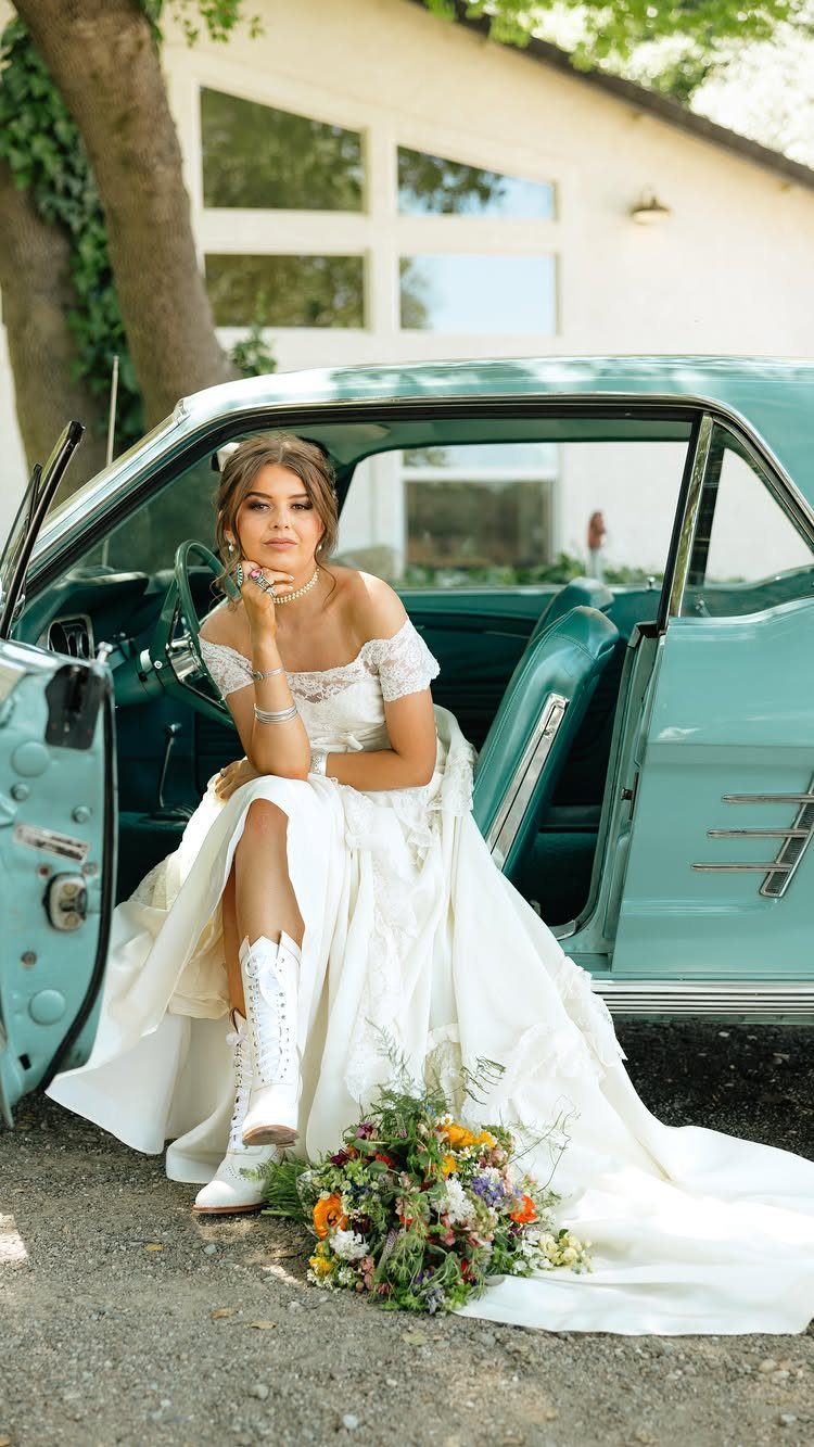Woman in a white lace wedding dress and cowboy boots by a teal car