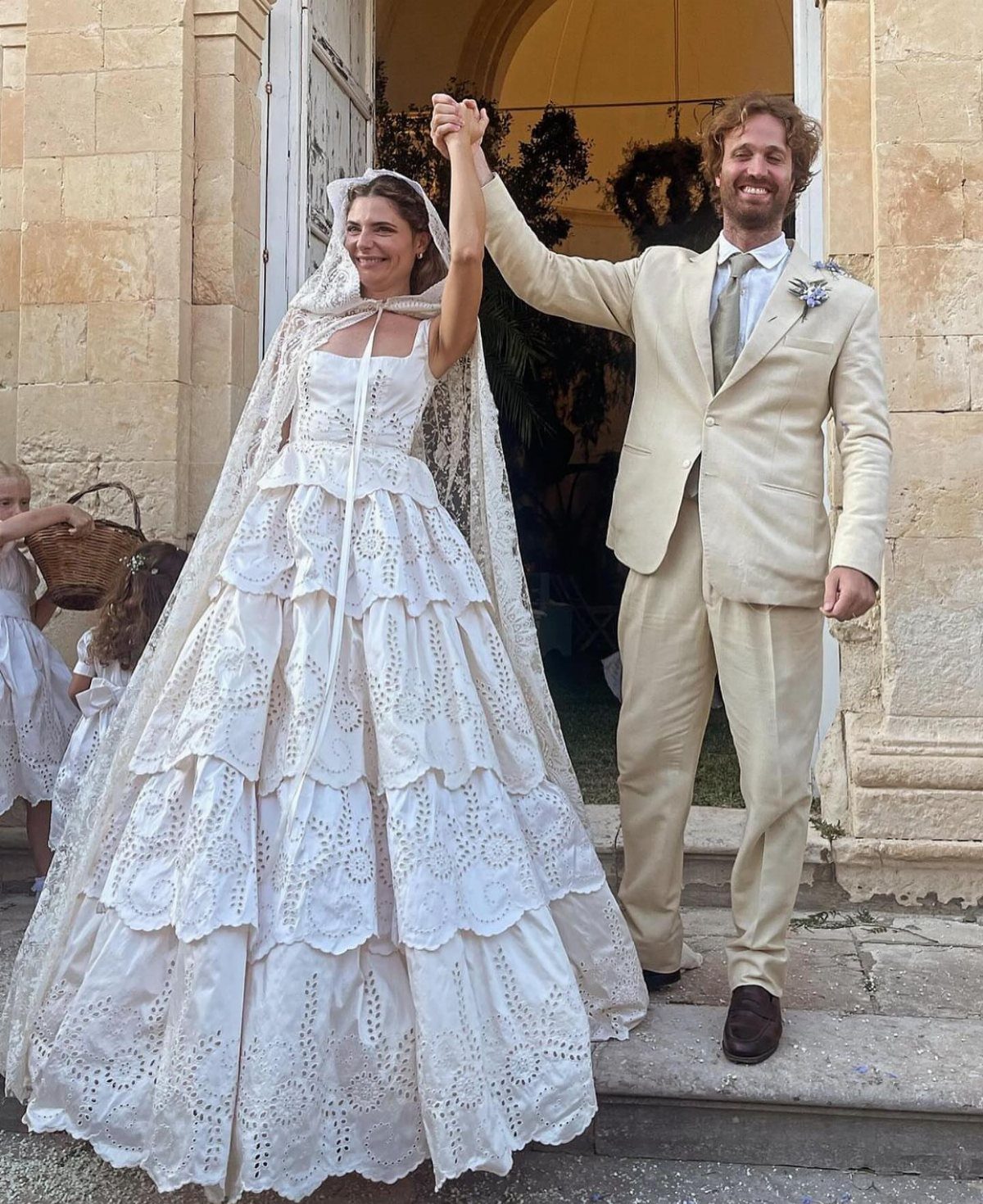 Bride in tiered white lace wedding dress and groom in beige suit holding hands.