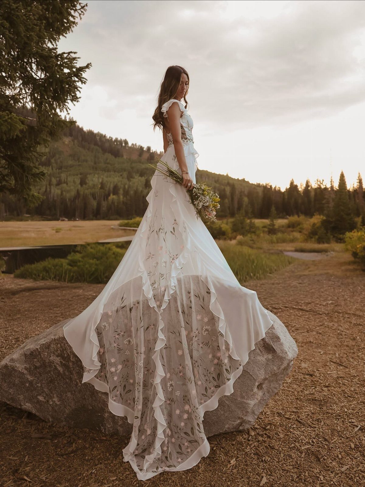 Woman in a white lace wedding dress standing on a rock in nature.