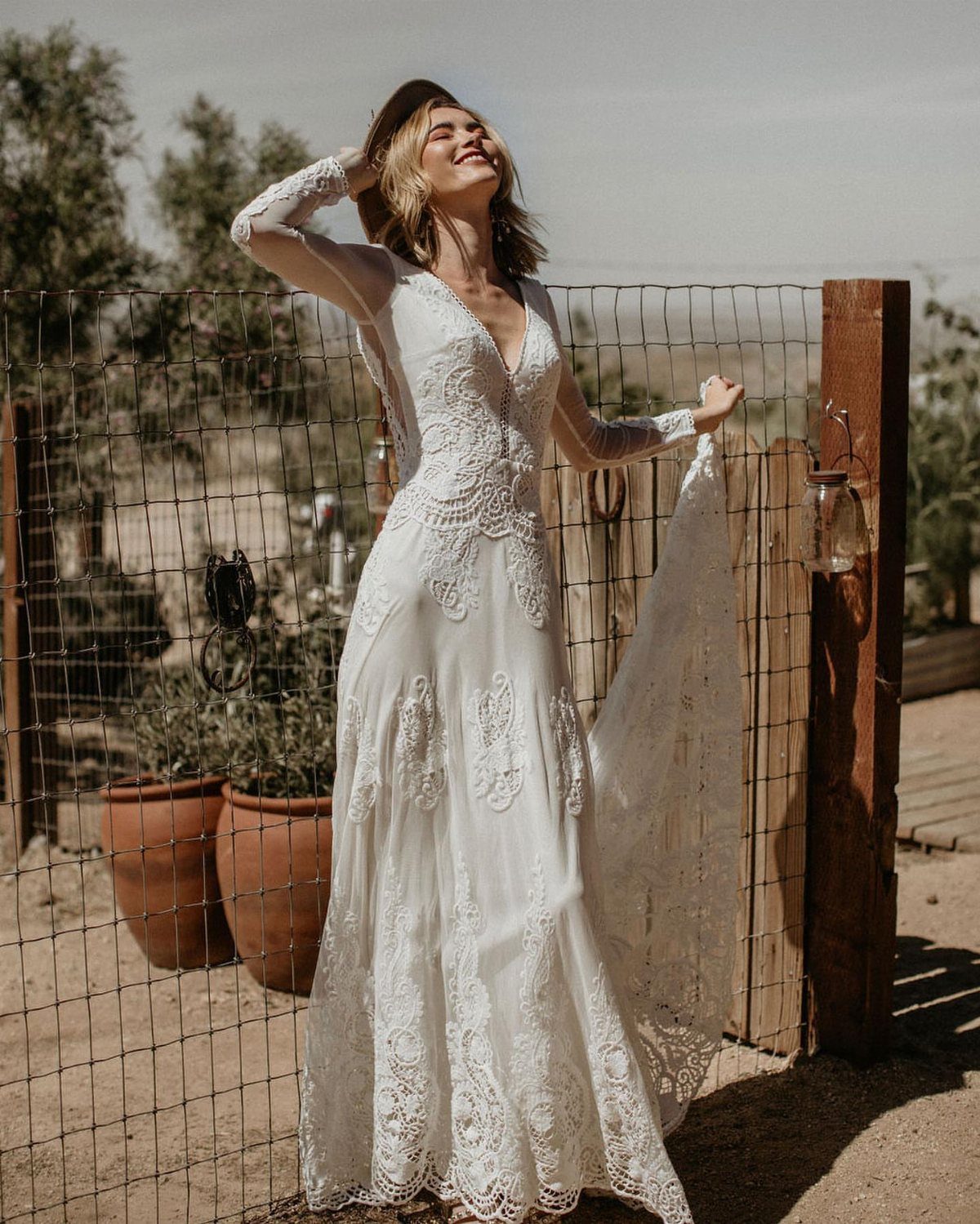 Woman in a white lace wedding dress and brown hat by a rustic fence.
