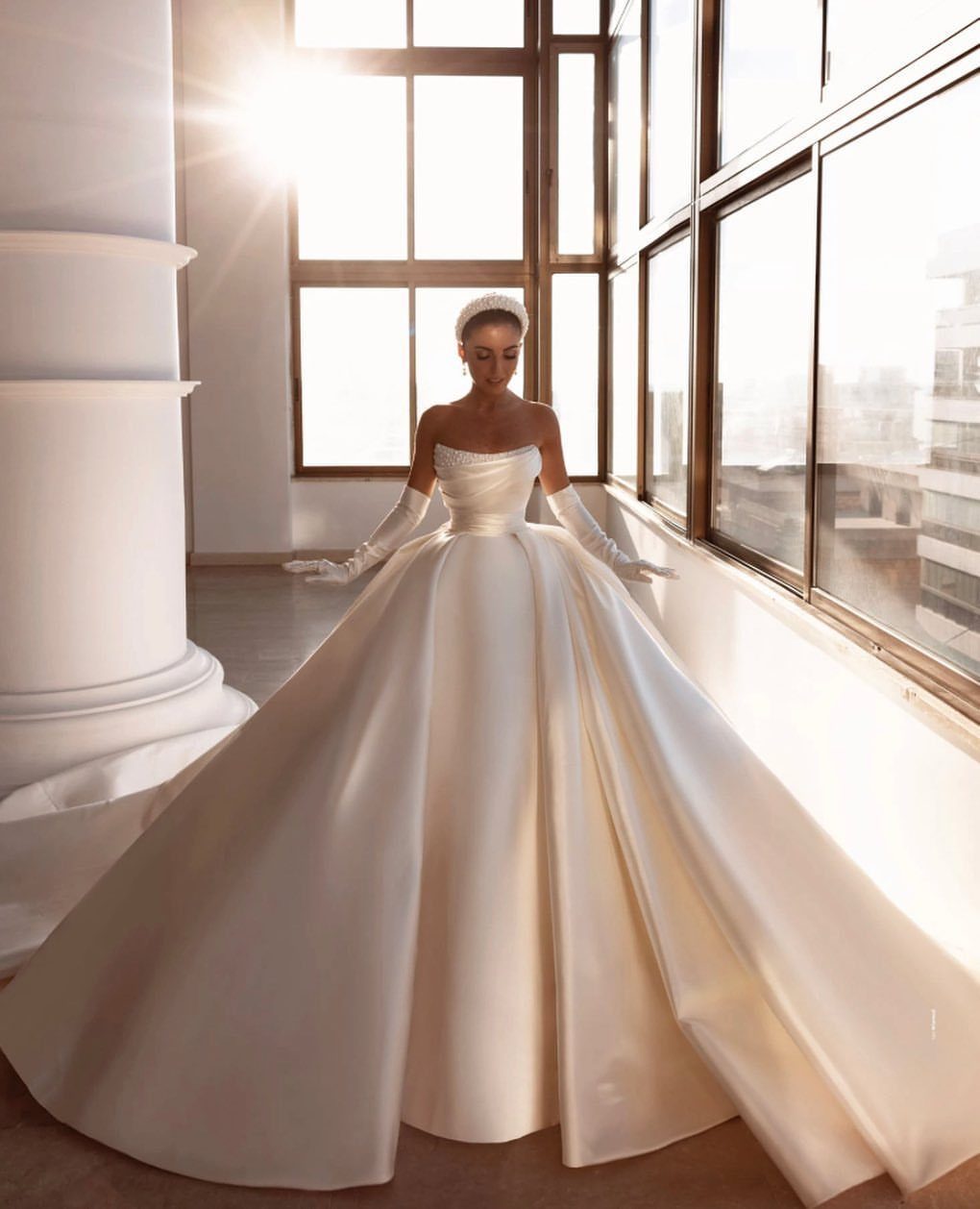 Woman in a voluminous white satin ballgown wedding dress standing by large windows.