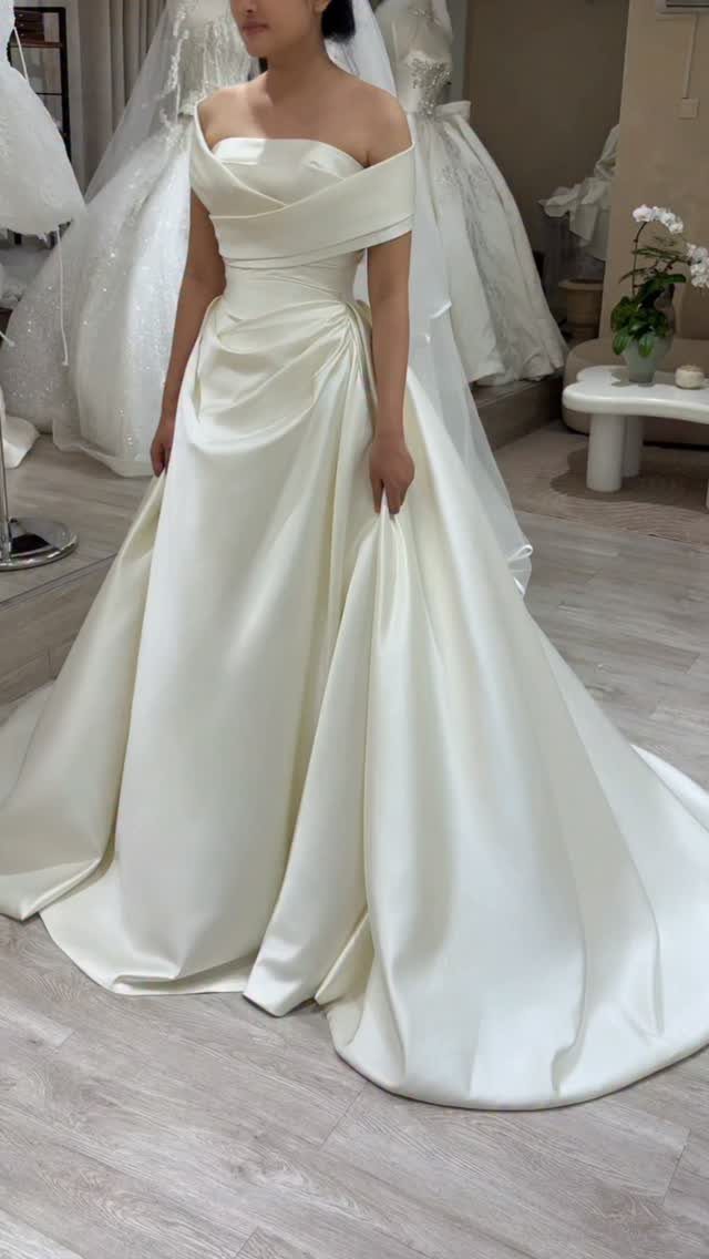 Woman wearing an off-the-shoulder white satin bridal gown in a bridal boutique setting.