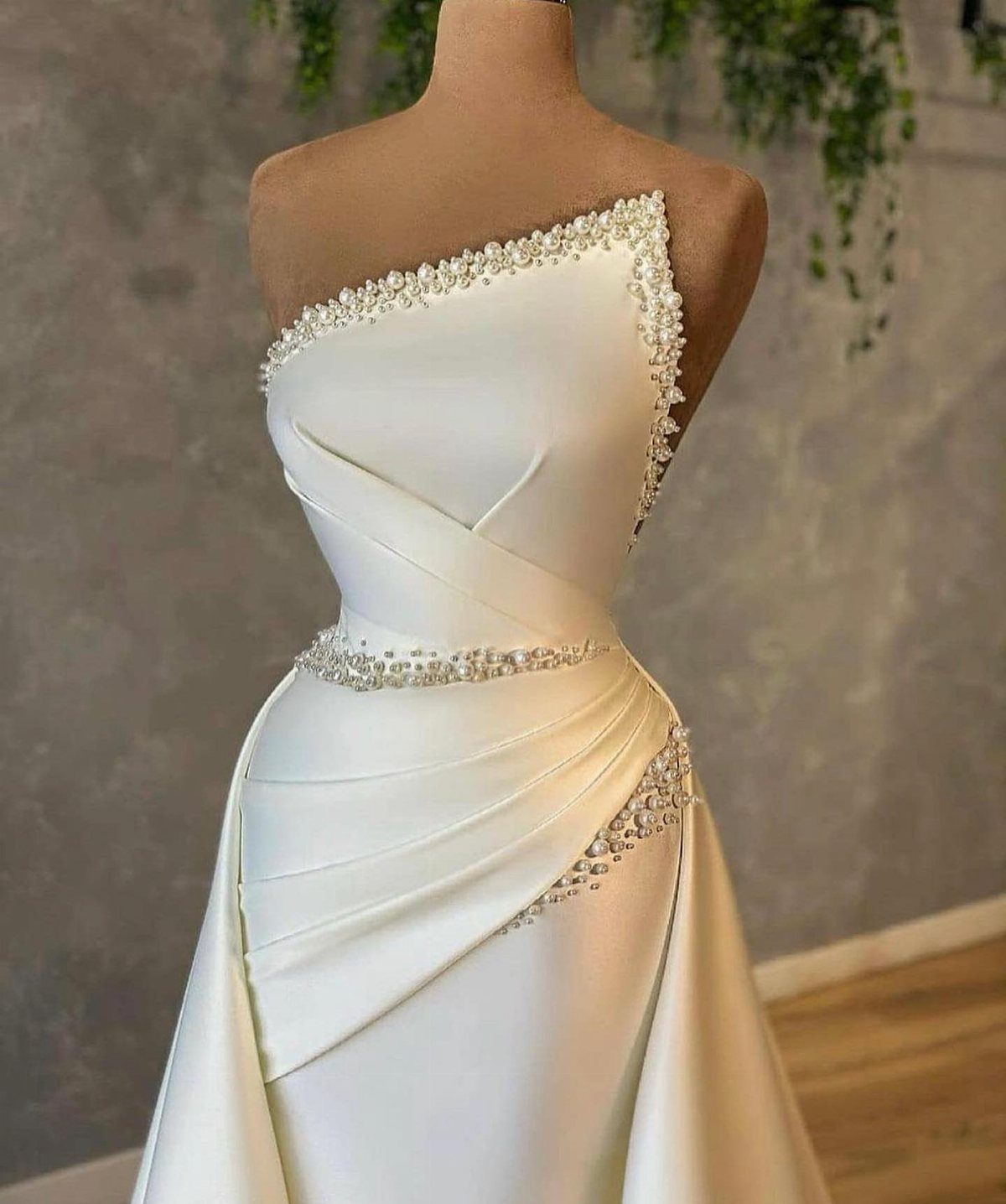 White satin bridal gown on a mannequin with one-shoulder pearl beading and draping.
