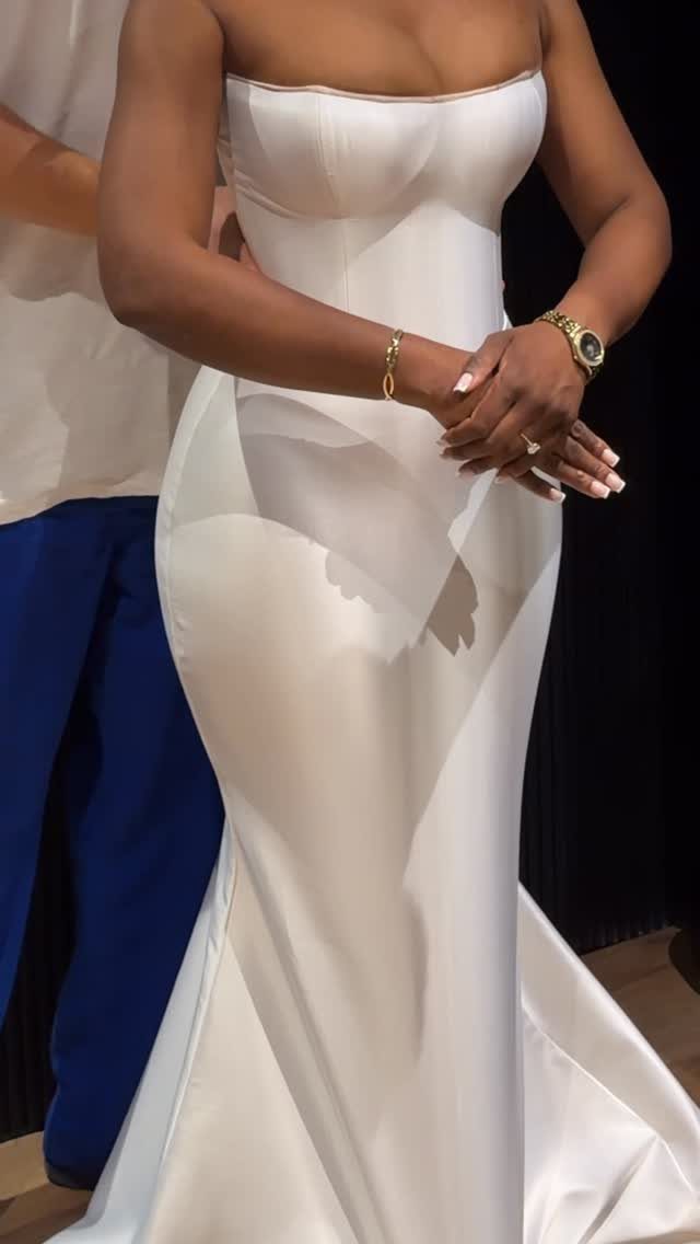 Woman wearing a strapless, form-fitting white satin bridal gown with gold jewelry.