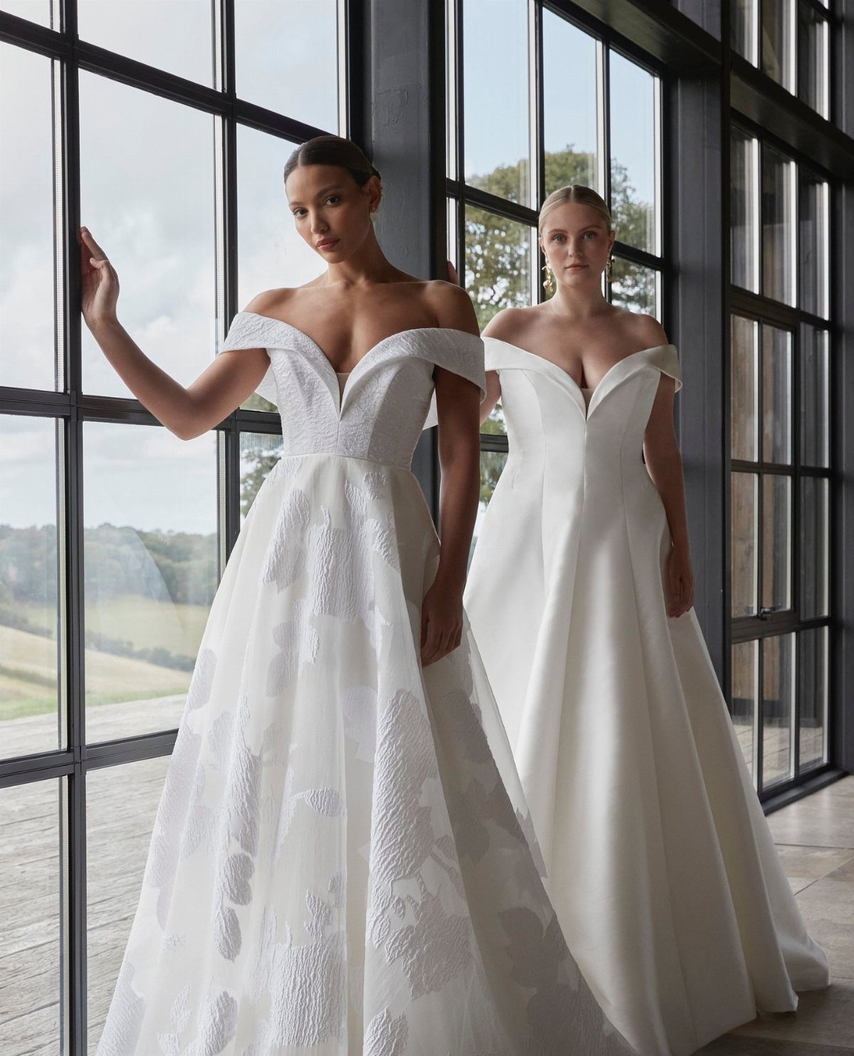 Two women in white bridal gowns posing by large black-framed windows.