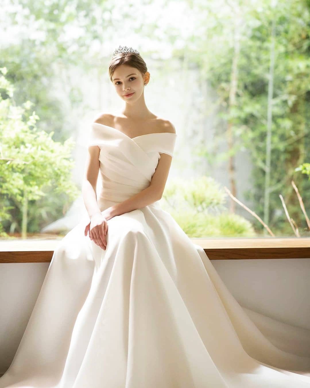 Woman in a white off-the-shoulder silk wedding gown sitting by a bright window