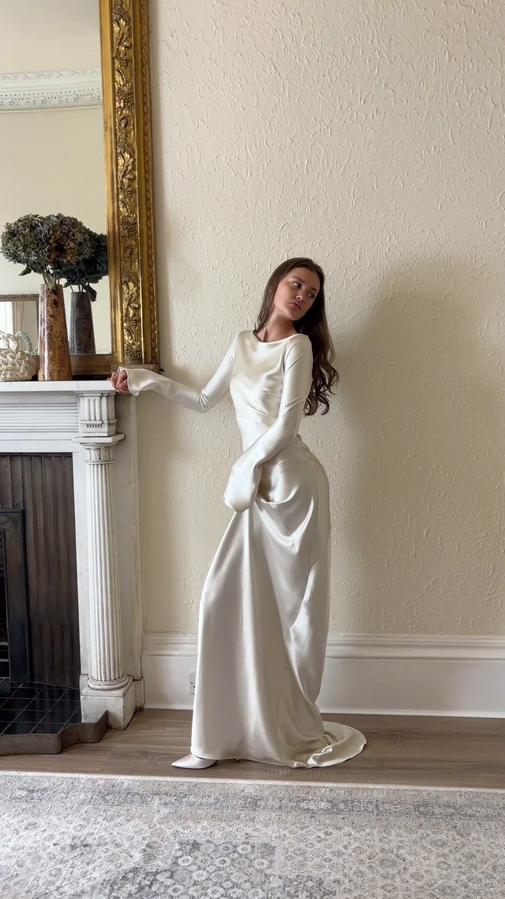 Woman in a long-sleeved white silk wedding gown posing by a fireplace.