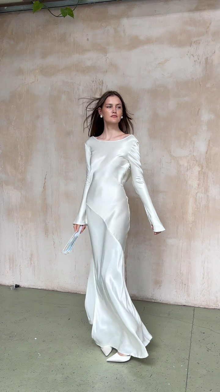 Woman in a long-sleeved white silk wedding gown standing against a textured wall.