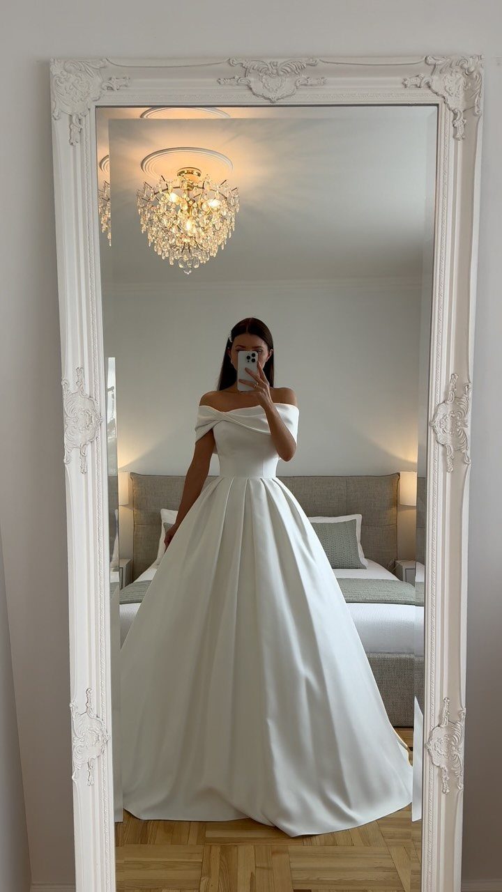 Woman in a white off-the-shoulder silk wedding gown posing in a mirror