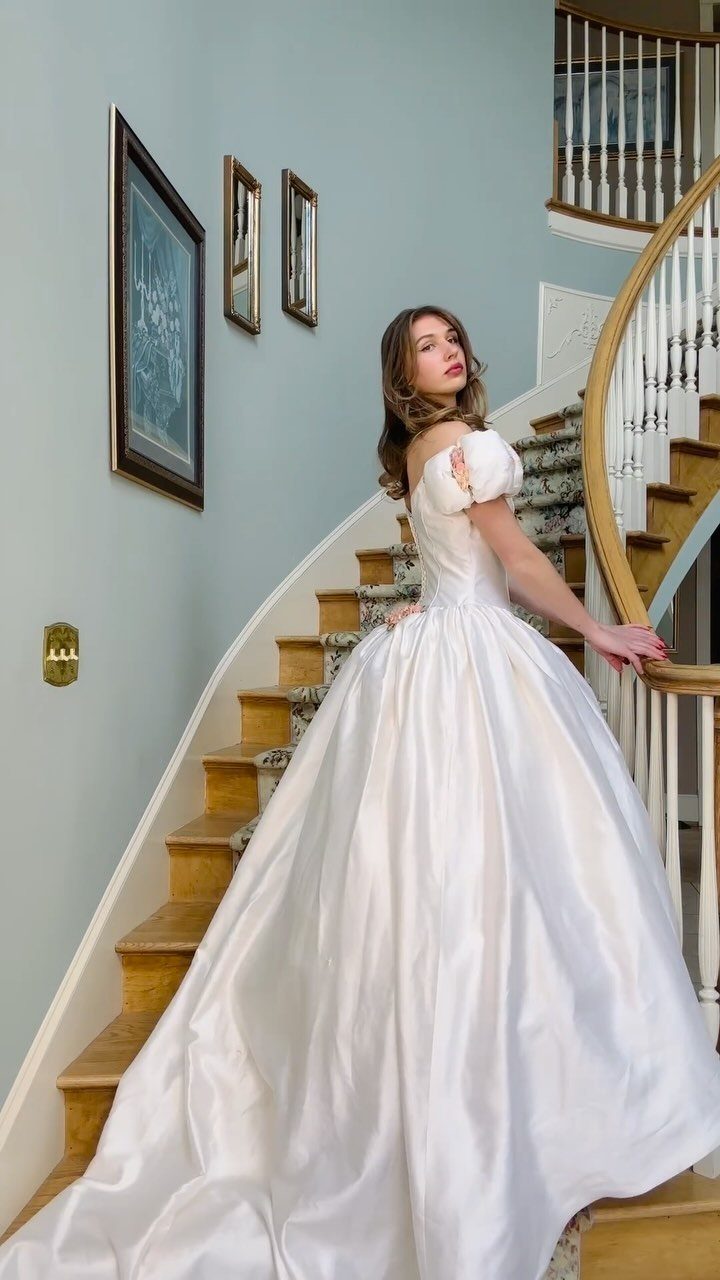 Woman in a voluminous white silk wedding gown standing on a wooden staircase.