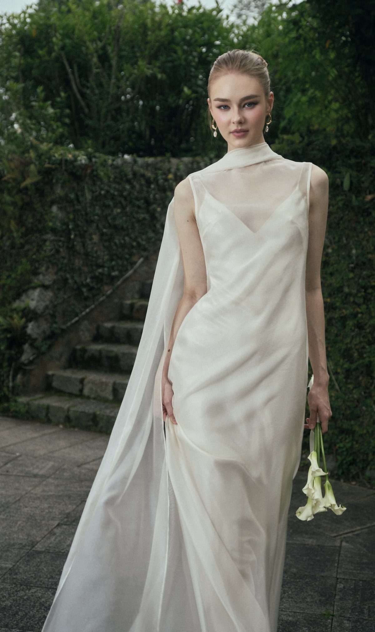 Woman in a white silk slip wedding dress with a sheer cape and lilies.