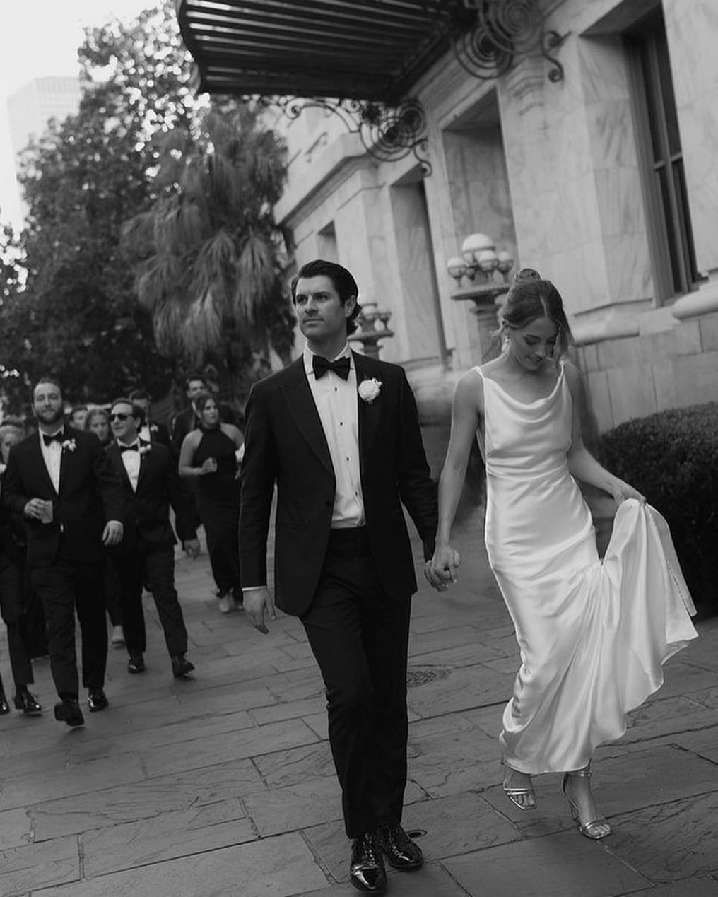 Bride in white silk slip wedding dress walking with groom in black tuxedo.