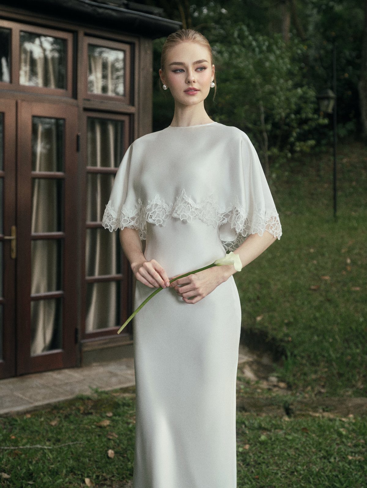 Woman in a white minimalist wedding dress with lace capelet holding a calla lily
