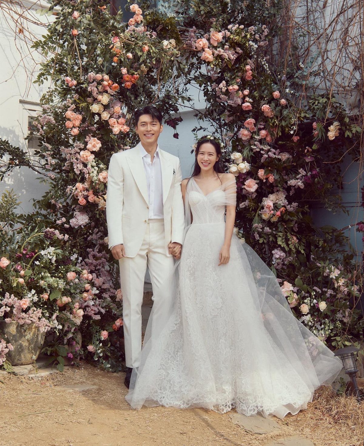 Couple in white wedding attire posing before a lush wall of pink flowers
