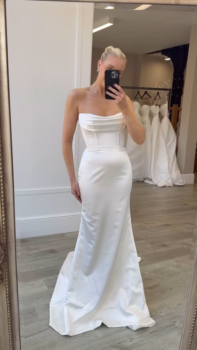 Woman in a white strapless satin wedding dress taking a mirror selfie in shop