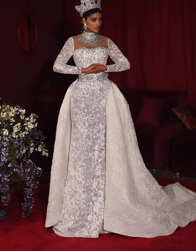 Woman in a white lace wedding gown with a crown and voluminous overskirt.