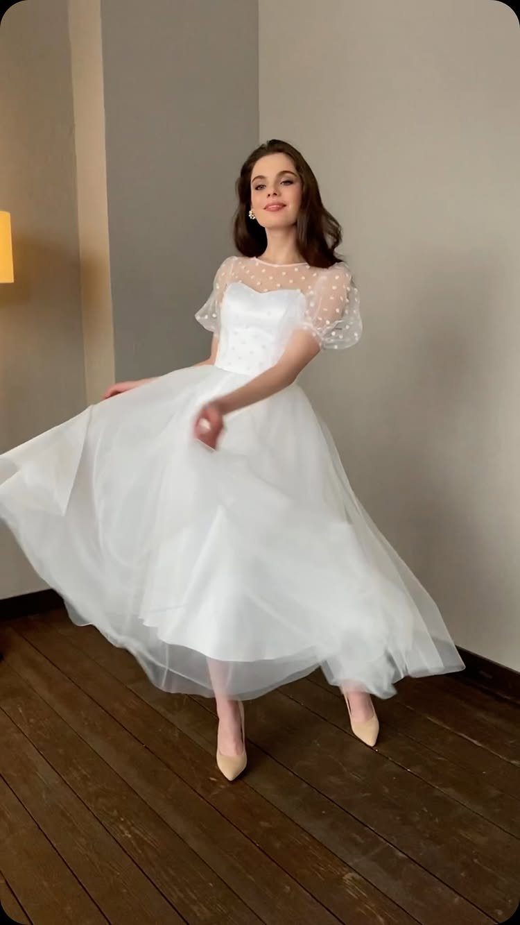 Woman in a white tea-length wedding dress with dotted sheer sleeves posing indoors.