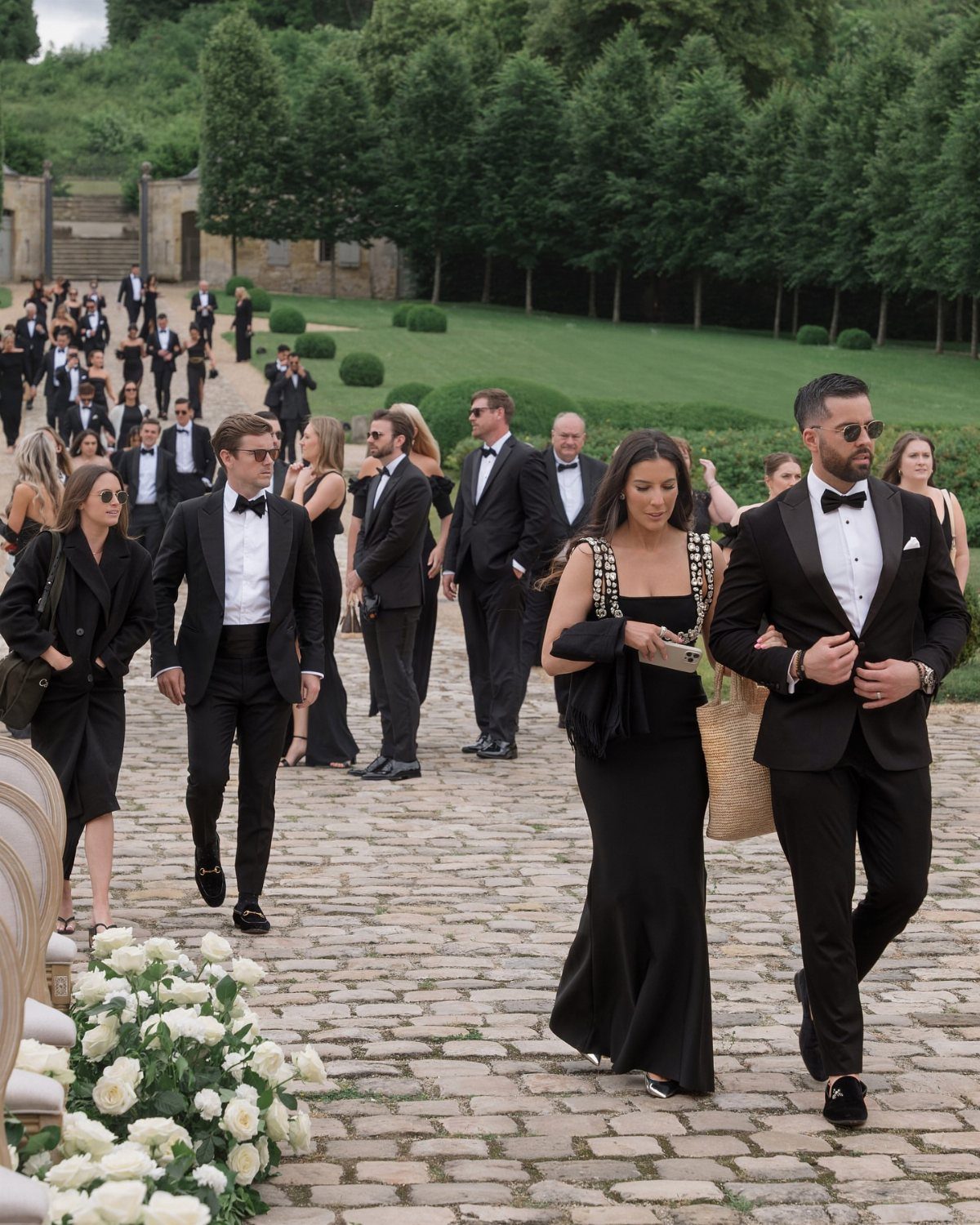 Guests in black tie attire walking on a cobblestone path at a wedding.
