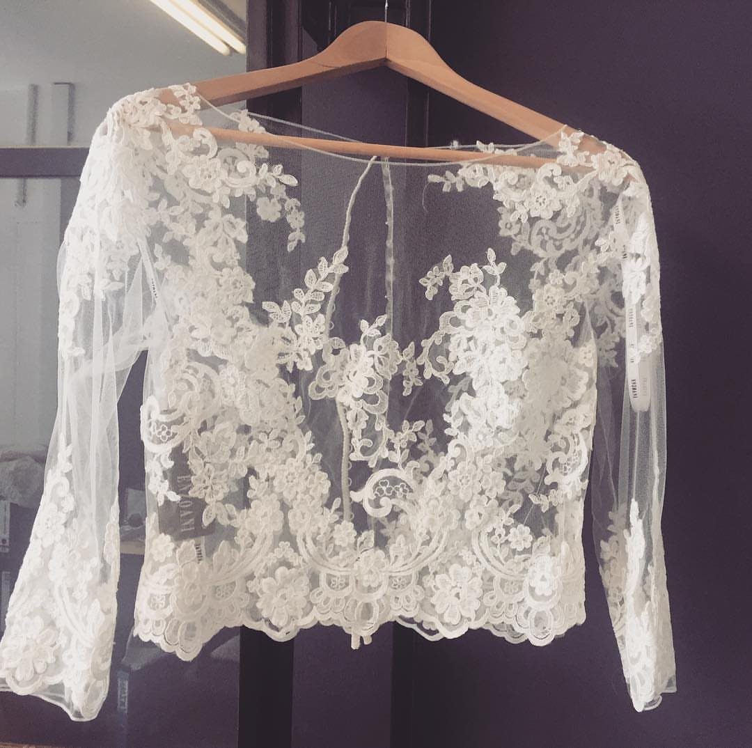 White lace bridal bolero with floral embroidery hanging on a wooden hanger.