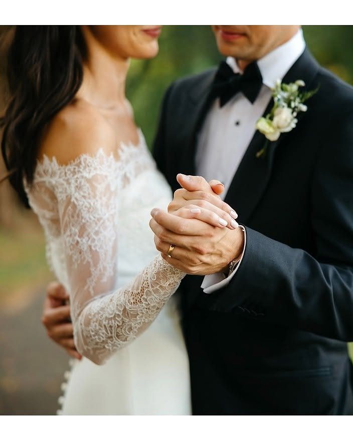 Bride in white lace bolero and groom in black tuxedo holding hands outdoors