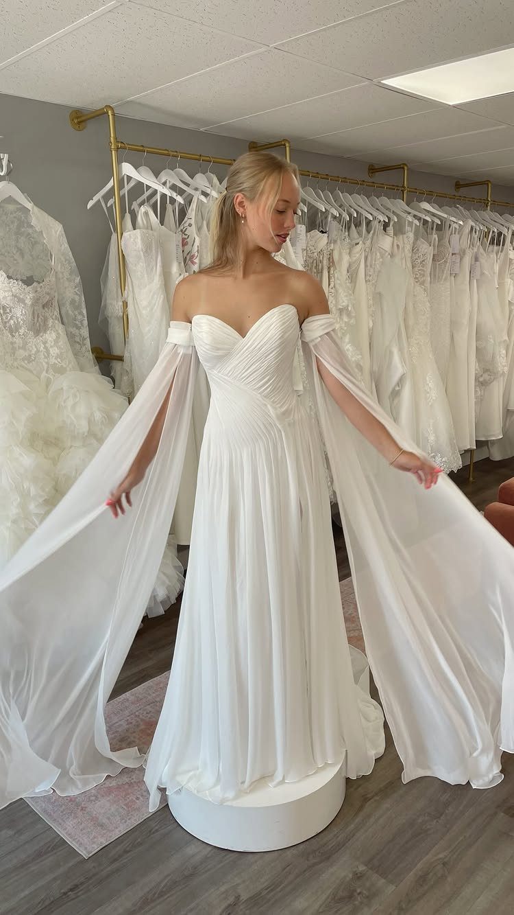 Woman in a white off-the-shoulder chiffon wedding dress with flowing cape sleeves.