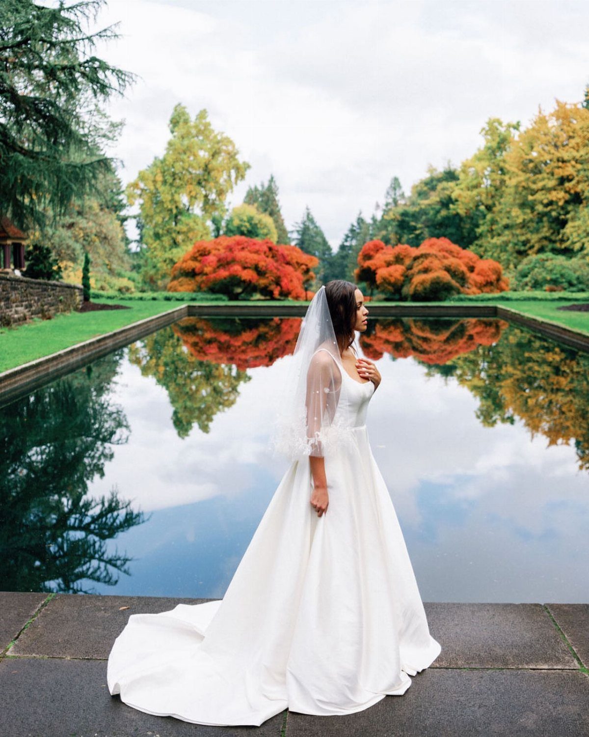 Bride in white wedding dress standing beside a pond with autumn foliage background